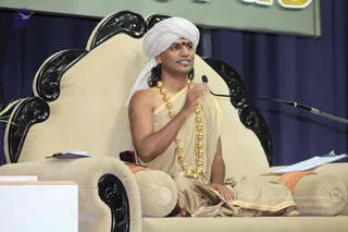A man seated on an ornate cream upholstered throne with black carved wooden armrests and back, speaking into a microphone on a stand. He wears a white turban with a red bindi on the forehead, a bare-chested upper body with a tan-gold dhoti and shawl, and a long necklace of large gold beads. His right hand rests at his chest while his left hand lies on his knee. Behind him hangs a blue curtain and a green banner. Clipboards and papers sit on the armrests and a small table stands at lower left. The setting suggests a discourse stage. Swamiji (probable) is shown in this discourse setting, identified by the page's album context and the distinctive turban, gold bead necklace and bare-chested saffron-toned attire, seated as the speaker.