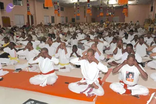A large indoor hall filled with seated people in white clothes performing a hand-over-eyes meditation posture. Many men with shaved heads sit on orange mats with red and yellow waist sashes, hands covering their closed eyes. Hanging colourful decorative banners and orange drapes decorate the ceiling. In the background, a distant figure appears near a small altar. The scene suggests a satsang or meditation session.