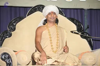 A seated man in a beige-gold sannyasi-style wrap and a white turban sits cross-legged on an ornate cream upholstered throne with black carved wooden arms. He wears a heavy gold bead necklace and a red bindi, and his hands rest on his lap. A microphone stand stands at the right against a purple draped backdrop, with orange and cream bolster cushions at the lower left. Appearance cues match a person seated in a discourse setting, consistent with the page's yes flag. The page excerpt is a discourse transcript on right listening.