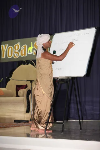 A man in a white turban and a gold-brown draped robe with a gold-bordered edge stands on a stage writing on a whiteboard on an easel. He holds a microphone in his other hand, and a gold beaded necklace hangs at his chest. The whiteboard grid contains handwritten words including Sattva, Rajas, Tamas, Peace, Restless, Silence and Energy. Behind him is a banner reading Yoga Sutra and a cream upholstered throne-style chair with a red cushion.