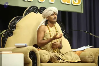 A man seated on an ornate cream-upholstered throne with carved black wooden armrests and back speaks at a microphone on a stand to his right. He wears a cream turban with a red bindi on his forehead, a saffron-gold wrap over his bare chest, and a long necklace of large gold beads. His right hand is raised in explanation while his left rests in his lap. A clipboard and a notepad sit beside him, a green banner with large gold lettering hangs above, and dark blue drapes form the backdrop. A printed copyright notice overlays the lower right of the photo.