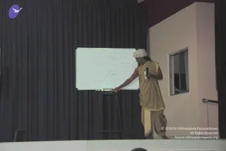 A man stands on a raised stage in front of a dark purple curtain, pointing at a whiteboard with a marker while holding a handheld microphone in his other hand. He wears a white turban, a beaded yellow-orange mala necklace, and a tan sleeveless top with gold trim over tan dhoti-style trousers. The whiteboard holds a handwritten grid of words with a few columns, and a small projector or lamp sits on a stand near the board.