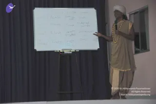 A man in a white turban and a tan-gold sleeveless tunic with matching draped dhoti stands on a stage beside a whiteboard on an easel, gesturing toward it with one open hand while holding a wireless microphone. A gold-beaded necklace hangs at his chest. The whiteboard shows a handwritten grid of Sanskrit and English terms in rows, including Satva, Rajas, Tamas, Peace, Silence, Sound, Dull, Depression, Chaos and Maya. A dark curtain forms the backdrop, with a window to the right. Black-dressed onlookers are not visible. The man appears to be giving a talk on the Yoga Sutras.