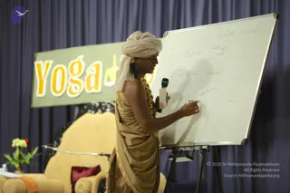 A man wearing a beige turban and a draped beige-gold robe with gold trim stands at a whiteboard, writing on it with a marker while holding a white handheld microphone. He wears a beaded necklace of golden-brown beads. The whiteboard grid contains handwritten words including 'Sattva/Rajas/Tamas', 'peace', 'Silence', 'Sound' and 'Irritate'. Behind him a green banner reads 'Yoga' in large yellow letters, and a cream upholstered chair with a red cushion and a small flower arrangement sits on the stage at left against navy drapery.