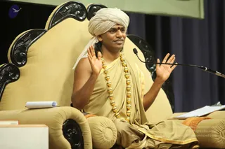 A photograph of a seated man on an ornate cream-upholstered throne-style chair with black carved wooden armrests, raising both open palms in an explanatory gesture while speaking. He wears a cream turban with a red tilak on his forehead, a beige-gold robe with gold trim, and a long strand of large gold beads around his neck. A microphone on a stand is at right, a notepad and clipboard sit on the armrests, and a dark blue curtain hangs behind him.