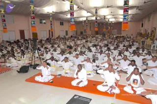 A large hall with pink walls and a white ceiling draped with fabric, decorated with tall colourful patterned pillars trimmed with orange tassels. Many rows of people in white clothes sit cross-legged on orange mats, many with yellow or red waist cords, performing a meditation with hands covering their ears and eyes. Camera tripods stand at the left edge. A black mat in the foreground reads Nithyananda Dhyanapeetam. Some people in the front wear white shirts with a printed portrait. Hands over the face and ears indicate a listening exercise.