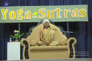 A man seated cross-legged on an ornate carved dark-wood and gold-upholstered throne-style armchair on a stage, hands folded in his lap. He wears a white turban, a gold-coloured draped robe with gold trim, and a long gold bead necklace. A microphone on a stand is beside him. Behind him hangs a green banner with large gold lettering reading 'Yoga Sutras' with a yellow yoga figure. A vase of red and white flowers and a water bottle stand on a white table to his left, in front of a dark blue curtain.