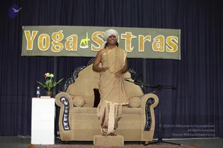 A photograph of a stage set for a Yoga Sutras session. A large banner reading "Yoga Sutras" in gold lettering with a yellow yoga-pose figure hangs above a dark blue draped curtain. A man stands on a low wooden step in front of a beige upholstered armchair with carved dark wooden arms and cushions. He wears a white turban, a beige draped robe with gold trim, and a long beaded necklace of gold-tone beads, with a red bindi on his forehead. He is smiling with his hands near his waist. A small white side table holds a glass water bottle and a vase of red and white flowers, and a microphone stand stands to the right.
