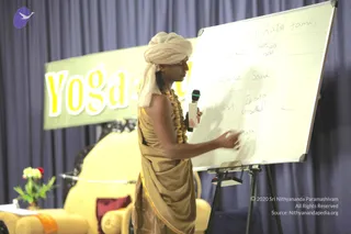 A man in a cream-colored turban and a tan draped robe with gold trim stands beside a large whiteboard, holding a handheld microphone in one hand and pointing with the other at handwritten notes in a grid. A yellow banner reading Yoga Sutra is behind him, with a cream-cushioned seat, a red and maroon cushion, and a small vase of flowers at the left. Swamiji (probable) is visible through the turban, tan robe and gold-trimmed shawl, and he is presenting a teaching on the board.