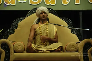 A man seated cross-legged on an ornate black-and-gold carved throne with tan velvet cushions, wearing a white turban, a tan and gold-bordered draped garment over one shoulder, and a long strand of large gold beads around his neck. He holds his right hand near his chest and faces forward toward a microphone on a stand. Behind him a green banner with large gold lettering reads partly visible text, with black drapery and a small potted plant at left.