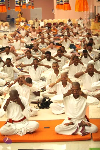 A large group of people in white clothes sits cross-legged on orange and striped mats in a hall with orange fabric hangings overhead. Many participants have their palms cupped over their closed eyes in a hand-over-eyes meditation posture, while a few sit with eyes shut and hands resting. Several men wear red cloth belts and white garments. A golden figure and seated devotees are visible at the back of the hall.