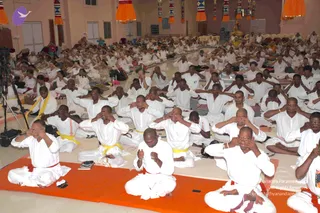 A large hall filled with many people seated on the floor in white ritual clothing, many with yellow or red waist cords, sitting on orange mats and white cushioned mats. Most participants sit with palms covering their eyes in a meditative practice, while a few in the front row sit with hands folded in prayer. Orange and multicoloured decorative hangings with tassels line the upper wall, and video recording equipment on a tripod stands at the left edge of the room.