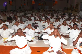 A large hall of seated participants in white clothes with orange and yellow waist sashes sits on orange and white mats. Most people, many with shaved heads, cover their eyes with their palms in a meditative listening practice. Hanging orange and red decorative tassels line the top of the room, and a small altar with ornate decorations is visible at the right edge. Headphones rest on the floor among the mats.
