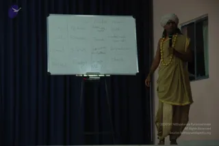 A man in a white turban and a golden-tan dhoti and shawl with gold trim stands at the right edge of a stage, holding a handheld microphone and wearing a long gold-bead necklace. Beside him stands a whiteboard on an easel in front of a dark blue curtain, showing a handwritten table with columns labelled Sattva, Rajas and Tamas, and rows for gross, subtle, primal and unmanifested, with words such as peace, silence, bliss, energy, restless, sound, depression and chaos. A small window is visible behind him on the right. The speaker appears to be giving a talk.