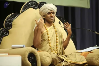 A man seated on an ornate cream upholstered throne with black carved wooden arms and a microphone on a stand before him. He wears a cream turban, a red bindi, a beige saffron-edged robe draped over one shoulder and a long necklace of gold beads. He gestures with both hands while speaking, and a notepad and clipboard rest beside him. Swamiji (probable): the appearance cues of a swept-back turban, ornate gold necklace and ceremonial robe are consistent with the page's attribution to Swamiji, seated in a discourse setting.