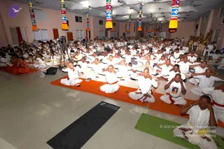 A large hall at a Nithyananda Dhyanapeetam-style yoga session with a crowd of several hundred people seated on orange and green mats in rows, mostly in white clothing with red or yellow waist cords. Many are covering their eyes with their hands in a meditative practice. A camera on a tripod films the group from the left. Colourful striped cloth pillars with yellow tassels hang from a white ceiling, and ceiling fans turn overhead. A black mat bears the printed text Nithyananda Dhyanapeetam. On the right, a seated figure in a white shirt with a printed photo sits near an ornate golden throne-like seat.