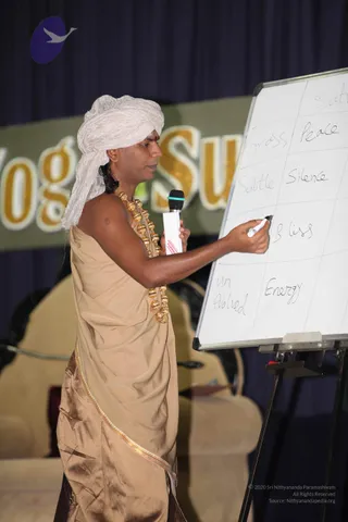 A man in a white turban and a beige draped garment with gold-brown border stands at a whiteboard on an easel, writing with a black marker. He holds a wireless microphone in his other hand and wears a long gold bead necklace of large gold-toned beads over his shoulder and chest. A red bindi is on his forehead. The whiteboard lists words such as Gross, Peace, Subtle, Silence, Bliss, Unevolved and Energy in a grid. Behind him a backdrop reads Yoga Sutra in large gold letters on a dark blue curtain, with an empty cream-coloured chair visible at left. He appears to be giving a talk at a Yoga Sutras session.