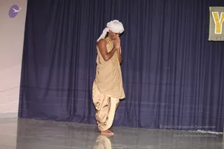 A single performer stands barefoot on a glossy grey stage floor in front of a deep navy blue curtain. He wears a white turban, a beige shawl draped over one shoulder, a gold-toned beaded necklace, and matching beige-gold dhoti-style trousers with a striped border. He stands with palms pressed together in anjali mudra at chest height, eyes lowered, in a respectful greeting pose. A gold letter Y is visible on a banner at the upper right, and a purple bird logo appears at the upper left.