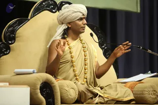 A photograph of a man seated on an ornate black-and-gold carved throne with a cream-coloured cushioned back and armrests. He wears a white turban, a tan-beige robe draped over one shoulder with a gold border, and a long necklace of large gold beads. A small red bindi marks his forehead. He gestures with both hands while speaking into a microphone on a stand; a notepad lies on the armrest and a clipboard sits to the right. A dark blue curtain forms the background. Swamiji (probable): long hair, turban and gold necklace on a throne-seated speaker giving a discourse.