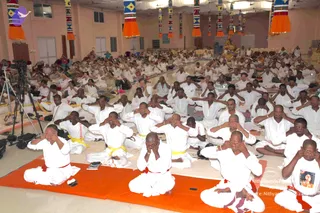A large hall filled with many seated people in white clothing, most wearing yellow or red waist and shoulder cords, sitting on orange mats and cushions. Many have their palms pressed over their closed eyes in a meditative hand-over-eyes practice. A tripod-mounted video camera stands at the left edge, and colourful woven panels with orange tassels hang from the pillars above. The hall has pale pink walls and white curtains, with more seated devotees visible toward the back.