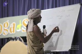 A photograph of a speaker at a whiteboard during a Yoga Sutras session. A man wearing a cream-coloured turban and a beige shawl draped over a tan garment, with a gold-toned beaded necklace, stands in profile holding a black microphone in one hand while writing with a marker in the other. The whiteboard is divided into a grid with handwritten words including 'Peace', 'Restlessness', 'Dull', 'Silence', 'Sound', 'Maya (delusion)', and 'Sattva', 'Rajas', 'Tamas' along the top. Behind him a banner reads 'Yoga Sutras' in orange and yellow letters beside a purple curtain. One person is visible.