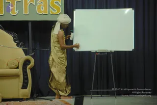 A photograph of a man in a gold-tan draped garment with a white turban and a yellow-and-orange flower garland writes on a white whiteboard on an easel. The whiteboard has faint handwritten words. Beside him is a gold upholstered armchair with carved black wooden armrests and an orange cushion, against a dark blue curtain. Above, a green banner with large gold letters reads 'tras' (Yoga Sutras session). The setting is a stage at a talk session. a person is named on the page but the man's appearance is not sufficiently distinctive to confirm, so he is described by role only.