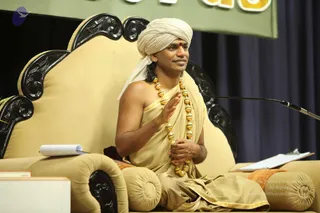 Photograph of a man seated on an ornate cream-upholstered throne with carved black wooden armrests and backrest, speaking at a microphone on a stage. He wears a cream turban, a plain beige-gold dhoti and shawl with gold border, and a long multi-strand necklace of gold beads. He has a red tilak on his forehead and holds his right hand raised in a teaching gesture while his left hand rests on his lap, holding a beaded item. A pale green banner with gold lettering hangs behind him, and a dark blue curtain is at the back. Papers and a clipboard rest on the throne. Swamiji (probable): the seated speaker wears a cream turban, gold beaded necklace and beige ceremonial shawl on the throne, cues consistent with the page's identification.