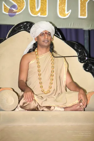 A seated man in a beige draped robe with a gold-edged shawl and a long gold bead necklace sits cross-legged on an ornate cream-upholstered throne with black carved wooden armrests. He wears a white turban, has a red bindi on his forehead, and has long dark hair falling behind his shoulders. A microphone stand is angled beside him. Behind him is a gold glittery banner with partially visible lettering, and a purple curtain hangs in the background. The figure appears to be Swamiji (probable), identified by the long hair, white turban, ornate gold necklace and beige saffron-family robes seated on a throne at a satsang-style discourse.