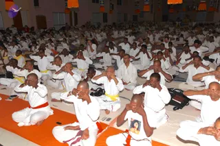 A large indoor hall filled with many seated people in white clothing, most wearing white dhotis or kurtas, with some having yellow or red cloth sashes at the waist. They sit on white floor mats and orange mats in rows, many with both hands raised to cover their closed eyes in a meditative practice. Orange and yellow hanging banners decorate the upper walls and ceiling. A few people in the front wear orange or red sashes and bead necklaces. No single leader is clearly visible in the frame, and no one can be identified as the a person from appearance here.