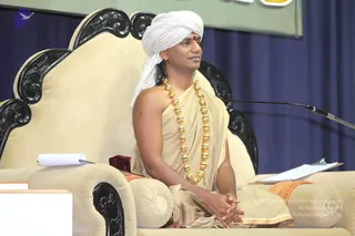 A man seated on an ornate cream upholstered throne with carved black wooden armrests and backrest, hands folded in his lap. He wears a white turban, a bare-shouldered beige and gold-bordered robe, and a long necklace of large gold beads. A red bindi marks his forehead. A microphone stand is at right and papers rest on the armrest, with a dark blue curtain behind and a framed picture above. Swamiji (probable), seated as the speaker, distinguished by the white turban, gold bead necklace and beige ceremonial robe.