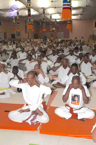 A large hall with white draped ceiling and colourful striped hanging banners with orange tassels. Many people in white clothes sit cross-legged on orange and cream mats, performing a hand-over-eyes meditation practice with palms covering their closed eyes. In the foreground a bald man in white kurta and orange-and-maroon dhoti sits in lotus-style posture, and beside him a man in a white T-shirt with a printed portrait of a woman in orange, wearing a rudraksha mala with an orange sash, does the same. A small deity murti or altar figure appears in the far background, with a few people in saffron-coloured clothing among the crowd.