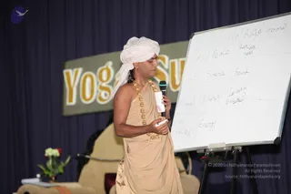 A man in a white turban and tan draped robe stands on a stage holding a wireless microphone in one hand and a marker in the other, facing a whiteboard. He wears a gold bead necklace. The whiteboard holds a handwritten table with headings Sattva, Rajas, Tamas and entries such as peace, restlessness, silence, sound, energy, and unsettled. Behind him a banner reads Yoga Sutras in gold letters on a dark blue curtain, with a small vase of white flowers on a side table. One person is visible.