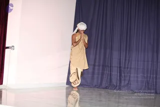 A single man stands barefoot on a glossy grey stage with his palms pressed together in namaste in front of his chest, head bowed. He wears a white turban, a beige-gold unstitched upper cloth draped over his shoulder and a matching beige-gold dhoti with a striped border, plus a beaded necklace of gold-toned beads. Behind him hangs a deep blue draped curtain, with a red curtain edge at left and a wall-mounted fixture. The floor reflects his figure. Swamiji (probable): the cues are the white turban, gold-toned beaded necklace and saffron-toned ceremonial dress, presented in a respectful greeting pose, so the appearance is only a plausible candidate.