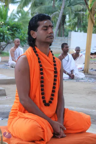 A man with long swept-back dark hair sits cross-legged with eyes closed in meditation on an orange cloth mat outdoors. He wears an orange saffron shawl and skirt, a long rudraksha mala reaching his waist, and a red bindi with white ash marks on his forehead. Behind him, three men in white dhotis sit on the ground under palm trees, one bare-chested with a sacred thread, and one with hands folded. A wooden pole stands at right.