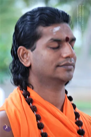 Close-up photograph of a man with eyes closed in meditation, head turned slightly to the side. He has long, swept-back dark hair, a red kumkum dot and white vibhuti marks on his forehead, and wears an orange saffron shawl over his shoulders with a dark rudraksha mala hanging across his chest. Blurred green foliage forms the background. The image is an appearance-based probable match for Swamiji based on the saffron clothing, rudraksha mala and swept-back hair, shown as a single person in meditative posture.