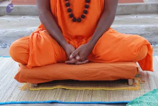 A seated figure in orange saffron robes sits cross-legged on a wooden platform with a woven straw mat beneath it, hands clasped in the lap in a meditative posture. A dark rudraksha mala with large beads hangs around the neck. The lower body and face are cropped out of frame; the setting is a pale stone floor with faint white decorative markings. The figure is a sannyasi-style devotee; identity is not established from appearance, so the a person field is unsure.