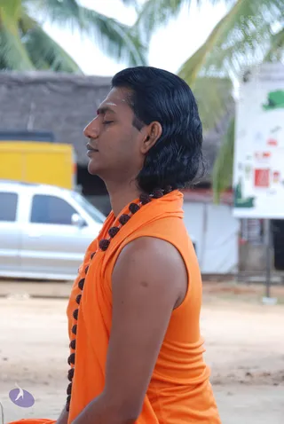 Side profile of a man seated outdoors in meditation with eyes closed, long swept-back dark hair, and a red tilak with white markings on his forehead. He wears a sleeveless bright orange garment with an orange shawl draped over his shoulder and a long brown rudraksha mala hanging across his chest. Behind him are palm trees, a thatched-roof structure, a silver car and a yellow vehicle, with a white board on the right displaying coloured graphics. A Swamiji (probable) candidate: long swept-back hair, orange clothing and rudraksha mala are visible cues, and the figure is physically present.