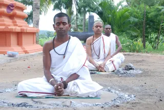 Three men sit cross-legged in meditation on bare earth outdoors, eyes closed, in plain white dhoti and shoulder cloths with red borders. The front man in the foreground wears a dark beaded mala and a bangle, hands folded in his lap; the two behind have shaved heads and tilak markings, one with a rudraksha-style mala. Behind them are an orange-painted shrine structure, a dark stone pillar, palm trees and green foliage. Grey ash or fire residue surrounds the seated figures, and a small plastic container sits at left.