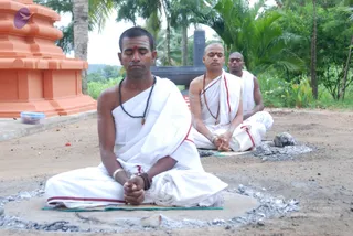 Outdoor photo of three men in white dhoti and shawl seated cross-legged in meditation on a sandy ground with ash-circle markings. The nearest man has eyes closed, hands clasped in his lap, a rudraksha mala and a red tilak on his forehead, and a gold bangle on his wrist. The middle man also meditates with a rudraksha mala and red tilak, and the man at the back sits with eyes closed. A tall orange stepped structure stands at left with palm trees and greenery behind, and a dark stone platform in the background. The scene is a meditation or padapuja session with no visible a person candidate matching the cues.