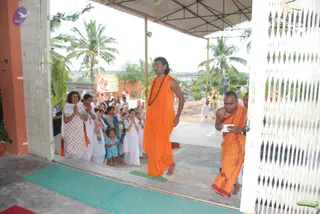 A photograph taken at an open-sided ashram entrance under a corrugated metal roof, with palm trees and a white latticed screen on the right. A tall man with long swept-back dark hair, bare-armed, wears a saffron-orange wrap and a long rudraksha mala, walking barefoot across a green mat toward the viewer. Beside him a shaved-headed monk in orange robes holds a folded white paper. Behind them a line of women in white and cream saris and salwar-kameez stands with palms pressed together in anjali, one small girl in a turquoise outfit among them. Further back, other white-clad devotees and orange-robed attendants move about a courtyard with stone statues and a red-and-yellow poster of deities on a wall. Swamiji (probable) is the tall man with long hair, saffron wrap and rudraksha mala, shown in person; this is a pada puja-style reception with devotees offering reverence.