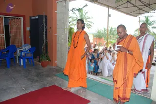 A photograph of a courtyard entrance at an ashram-style building with orange walls, a tin roof, palm trees and a red-and-green mat on the ground. In the foreground, a barefoot man with long swept-back hair and a rudraksha mala wears a sleeveless saffron-orange robe and walks forward. Beside him, a shaved-headed man in orange robes with a maroon border holds a small white card or paper while glancing down. At the right, a man in a white dhoti and shawl with a sacred thread stands holding an orange-handled item. Behind them, a crowd of devotees in white and blue clothes, some with hands folded in namaskar, stands watching near the doorway, with a child in a teal top among them. Blue plastic chairs sit to the left.