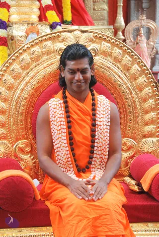 A seated man in saffron-orange robes sits on an ornate gold throne with a red cushioned seat and red bolsters. He wears a long dark rudraksha mala and a white and orange beaded garland, with hands folded in his lap and a red tilak on his forehead. Behind him are garlands of red, yellow and purple flowers and a gold-coloured ceremonial setting. A small scene with a shrine figure is visible at the upper right.