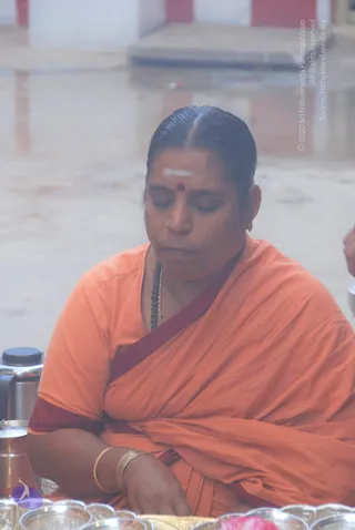 Photo of a woman seated cross-legged with eyes closed, wearing an orange sari with a maroon border and a maroon blouse, a black beaded mala around her neck, gold bangles on her wrist, a white vertical tilak and red bindi on her forehead, and a gold nose stud. In front of her stand polished silver vessels and a steel flask, with a pink flower visible at the bottom edge. The background shows a wet courtyard with red and white walls.