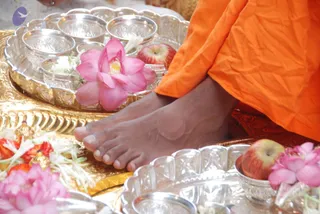Close view of bare feet of a seated person in orange robes resting on a gold tray during a padapuja foot-worship ritual. Silver bowls, pink lotus flowers, red apples, white flower petals and marigold garlands surround the feet on silver and gold trays. No face is visible, and no one else is visibly present in the frame.