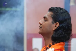 Close side-profile photograph of a man with long, swept-back dark hair and closed eyes, wearing an orange-saffron garment and a rudraksha mala with a white floral garland. Behind him is a pink and red wall with a blue-grey smoky background. The posture appears calm and meditative.