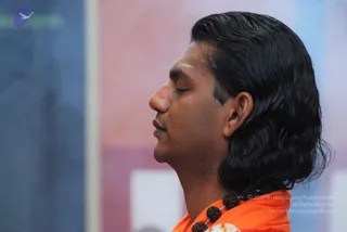 Side profile close-up of a man with long, swept-back black hair and eyes closed, wearing an orange garment and a dark rudraksha mala around his neck. A white vertical mark is visible on his forehead. The blurred background shows pink, blue and white panels.