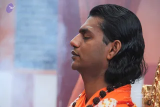 Close side profile of a man with eyes closed, seated in meditation before a soft pink and blue backdrop. He has long, swept-back black hair, a red tilak on his forehead and a white marking above it, and wears an orange-saffron shawl with a rudraksha mala of dark brown beads and white-and-red flower garland. A gold-coloured ornate chair edge is visible at the right.