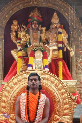 A seated man with closed eyes, long dark hair swept back, a red vibhuti or kumkum mark on his forehead, wearing an orange saffron cloth, a white flower garland and a dark rudraksha mala, sits on an ornate gilded throne. Behind him, a golden deity murti with a crown and multiple arms, adorned with yellow, red and purple flower garlands, stands beside a second golden figure in a red-draped niche with a gilded arch frame. The man is meditating in a temple setting. Probable Swamiji, identified by long swept-back hair, saffron clothing, a rudraksha mala and a ceremonial garland.