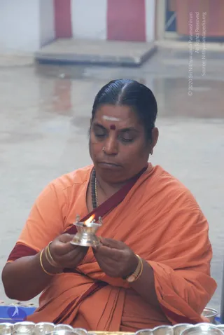 A woman in orange and maroon sari-style robes sits on a wet floor, holding a small silver oil lamp with a lit flame in both hands. She wears a white vertical tilak and a red kumkum dot on her forehead, a dark rudraksha mala, gold bangles and a nose stud. Silver offering plates sit in the foreground.