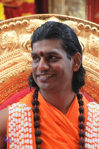 Close photo of a smiling man with long, swept-back dark hair, a red kumkum dot and white tilak on his forehead, wearing a saffron-orange shawl, dark rudraksha mala beads and a white-and-orange beaded garland. He sits in front of an ornate gold throne with repoussé floral carving and gold knobs. Pada puja setting.